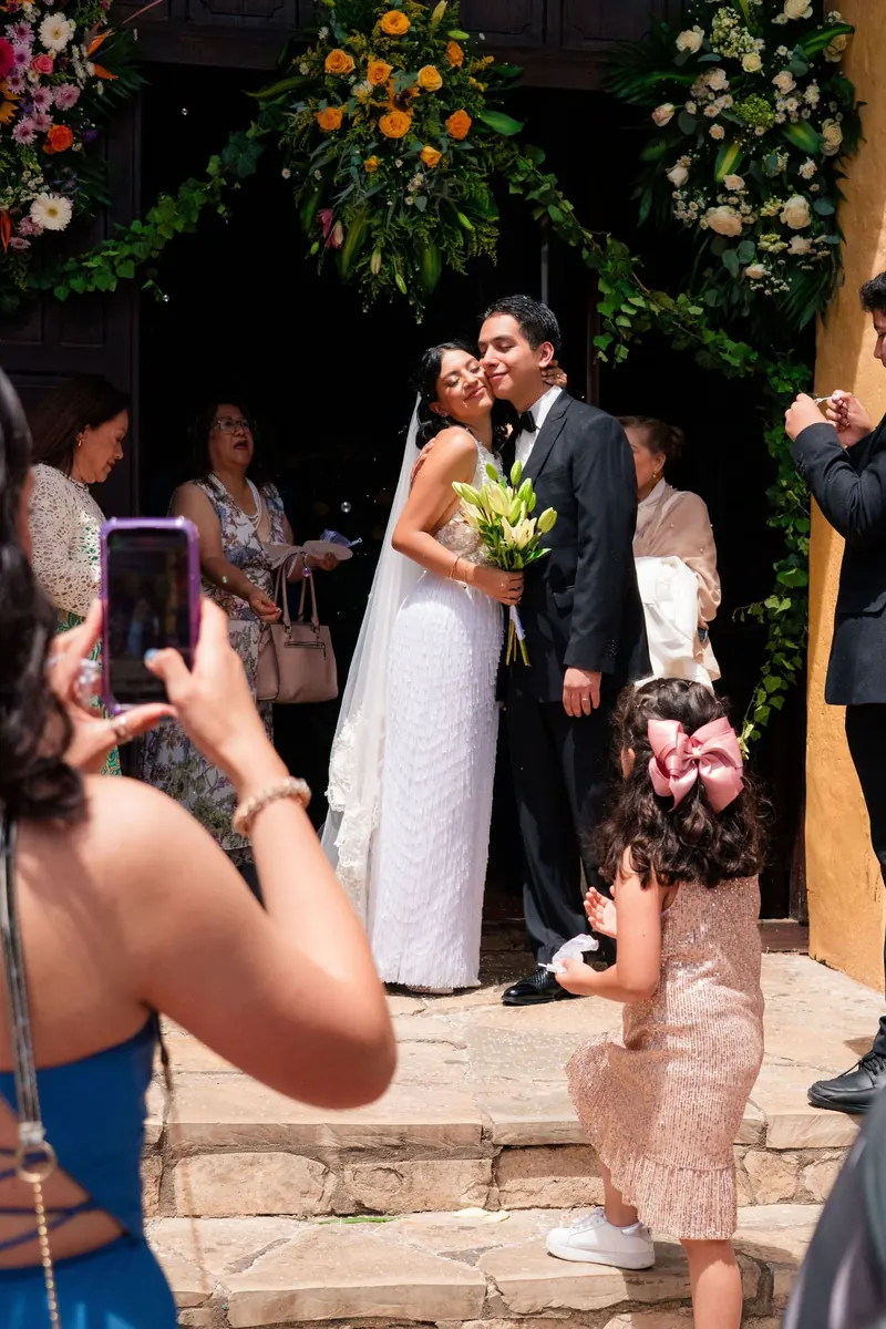 Newlyweds embracing under a floral arch at the church as a guest takes a photo