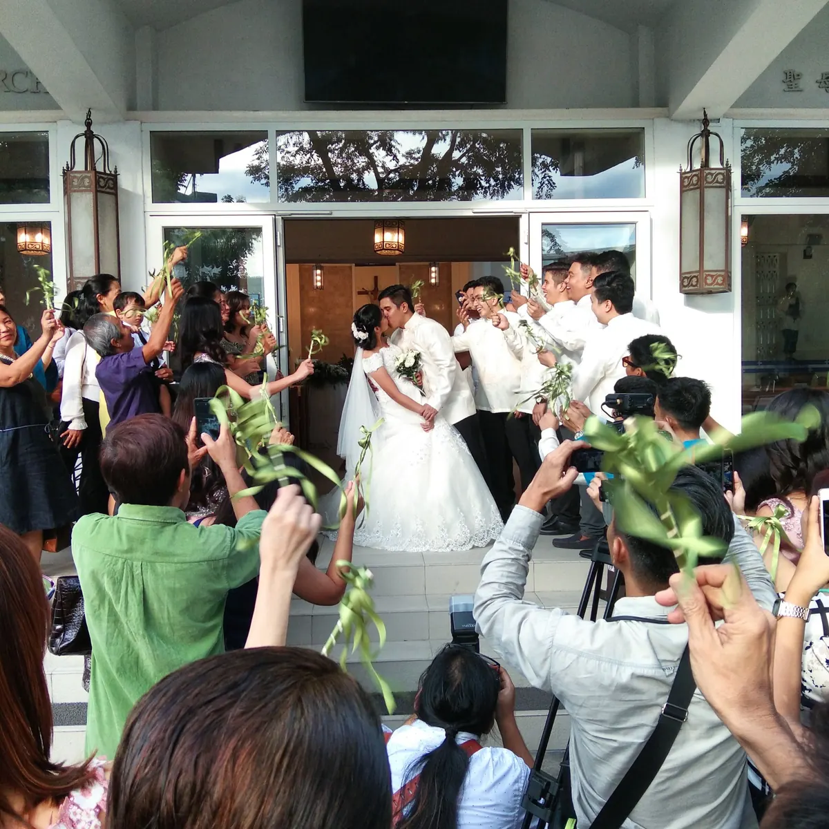 A couple kissing at the church doors while guests wave palm ribbons and film